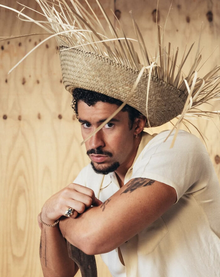 Man wearing a large woven straw hat, leaning on a wooden object, with tattoos visible on his arms indoors.