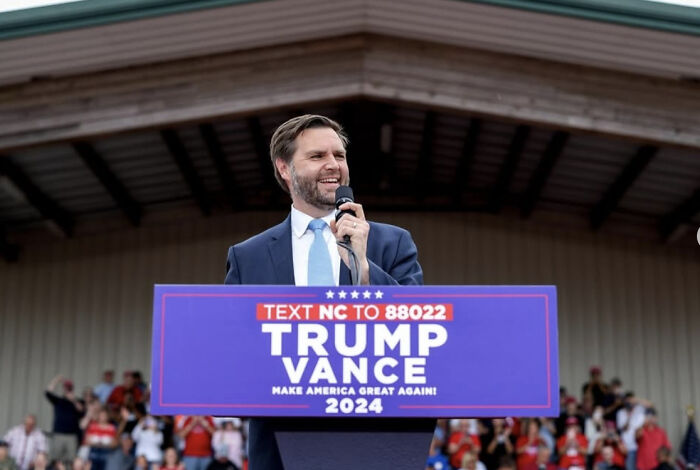 Man speaking at a rally with a Trump Vance 2024 sign, related to Gavin Newsom welcoming JD Vance in California.