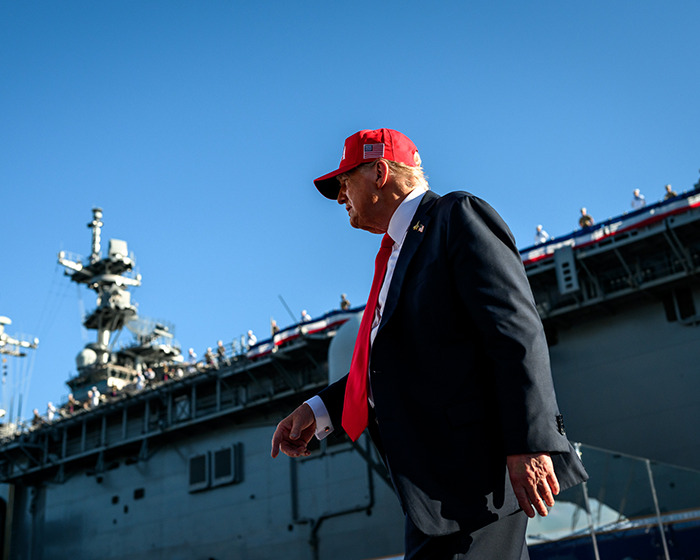 Former President Trump in a red cap walking near a military ship amid Venezuela war and CIA operations buildup.