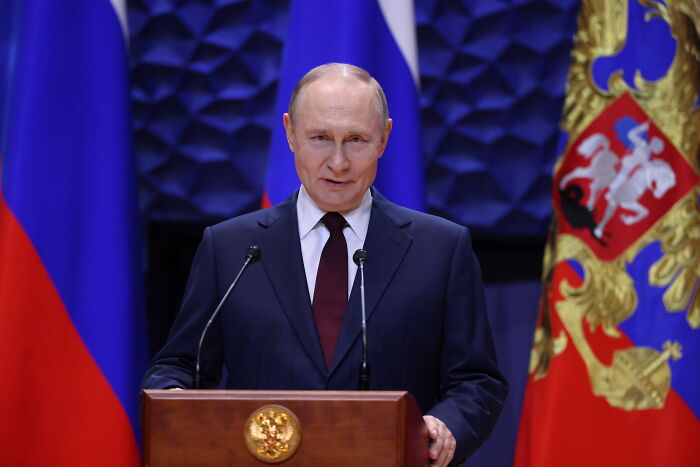 Man in dark suit speaking at podium with Russian flags behind him during a formal address session.