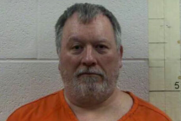 Tennessee man in orange jail uniform with gray hair and beard, detained over Trump meme posted in vigil group.