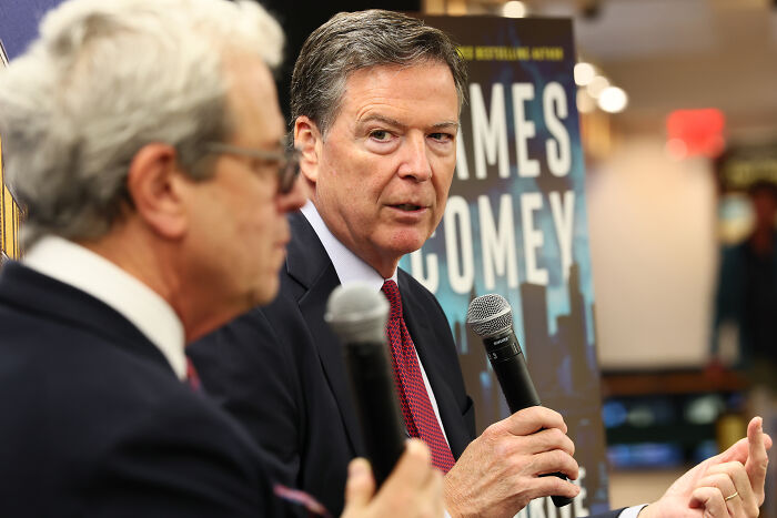 Two men in suits speaking at an event, with a James Comey sign visible behind them discussing FBI and ADL ties.