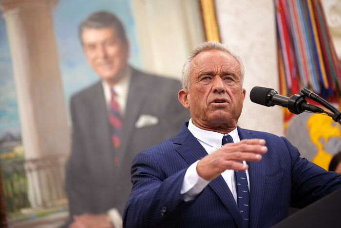 Man in a blue suit speaking at a microphone with a blurred portrait in the background, related to MAGA fake poster news.