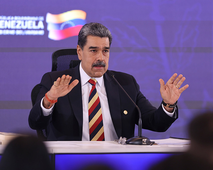 Venezuelan leader speaking at a podium with Venezuela flag backdrop amid warnings of war and military buildup.
