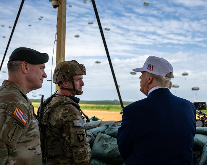 Donald Trump wearing a white hat with American flag talking to U.S. soldiers during a military parachute exercise.