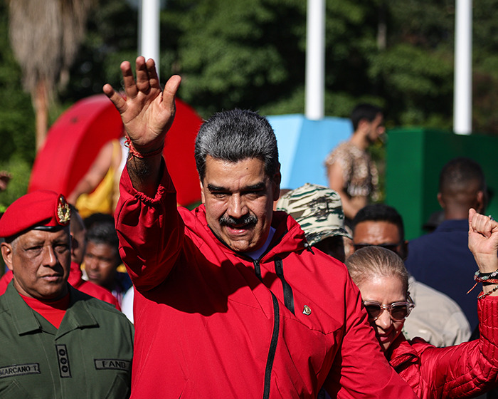Nicolas Maduro in a red jacket waving to a crowd with military personnel nearby during a public event.