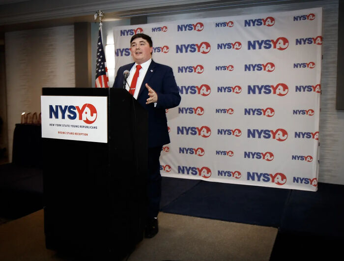 Young Republican leader speaking at podium with NYSYR backdrop, related to racist remarks in private chat report.