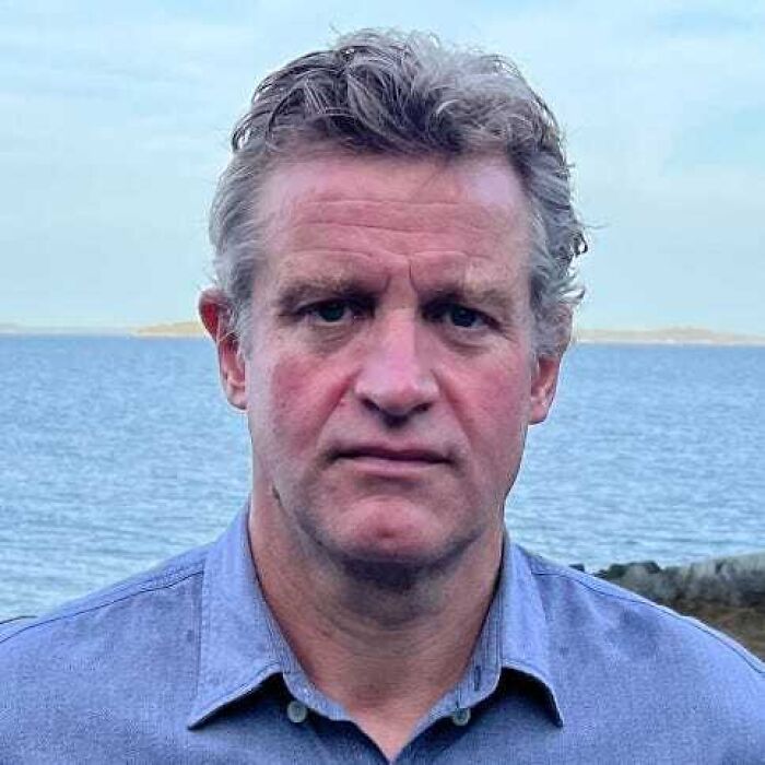 Middle-aged man with curly hair wearing a blue shirt, standing by the sea, related to influential people in Epstein&rsquo;s emails.