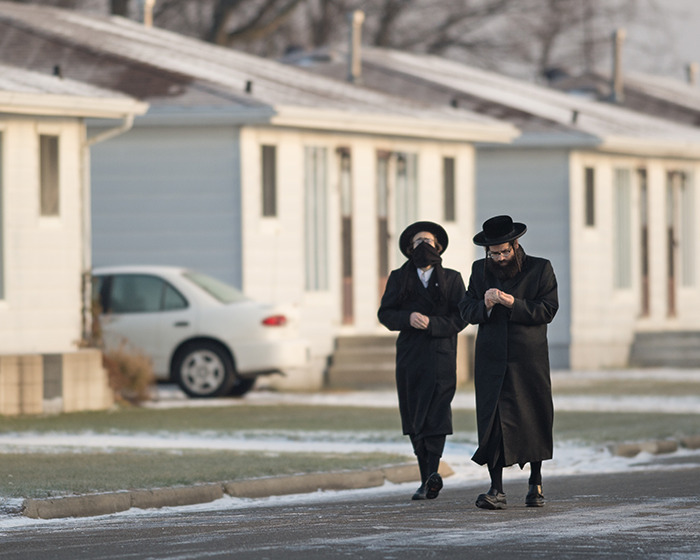 Two men in traditional Lev Tahor sect clothing walk near residential buildings on a cold day during police rescue efforts.