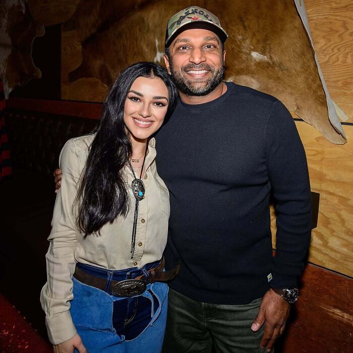 Kash Patel smiling with girlfriend at indoor event, wearing casual outfits with wood panel background.