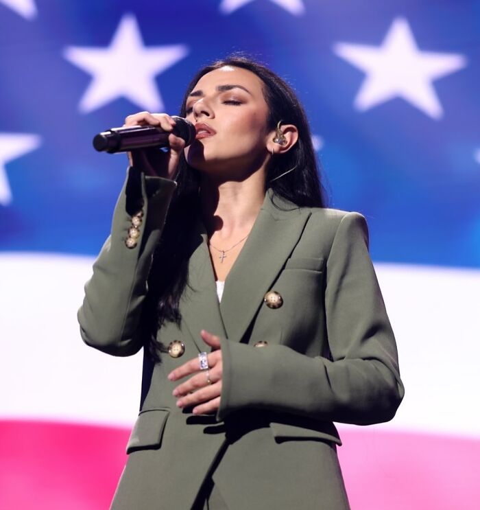 Woman singing on stage with microphone in front of American flag backdrop at a public event with Kash Patel theme.