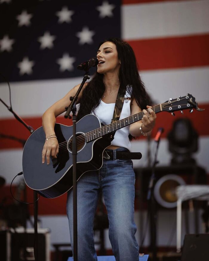 Female country musician performing on stage with guitar in front of an American flag, highlighting FBI Director Kash Patel news.