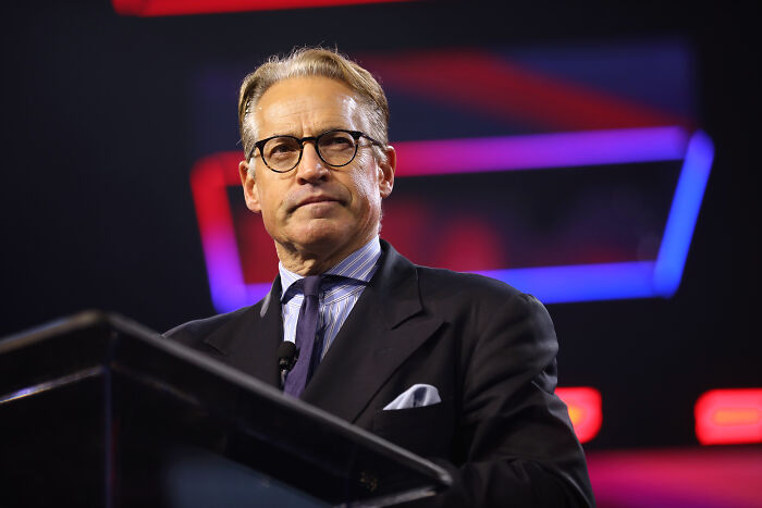 Eric Metaxas speaking at a podium wearing glasses and a suit, reacting to Trump crashing Mar-a-Lago wedding event.
