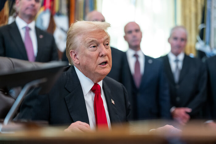Donald Trump speaking at a desk during a meeting with aides in the background, related to MAGA voter grocery prices.