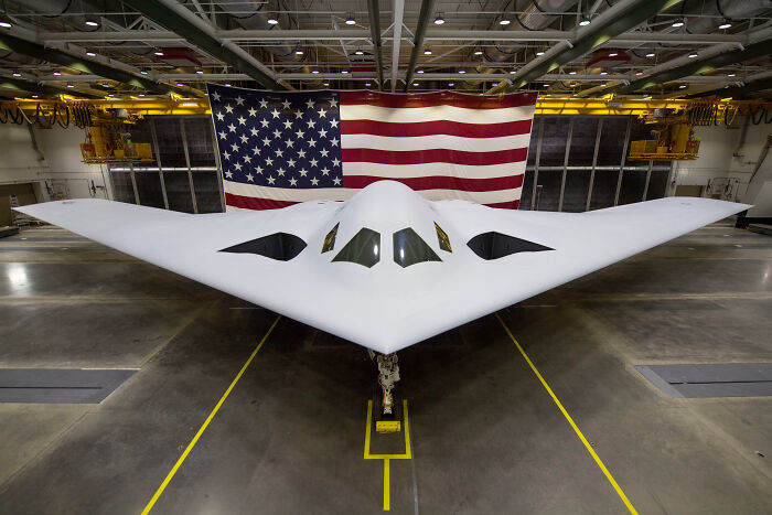 Stealth military aircraft in a hangar with a large American flag backdrop, symbolizing a dystopian troops address.