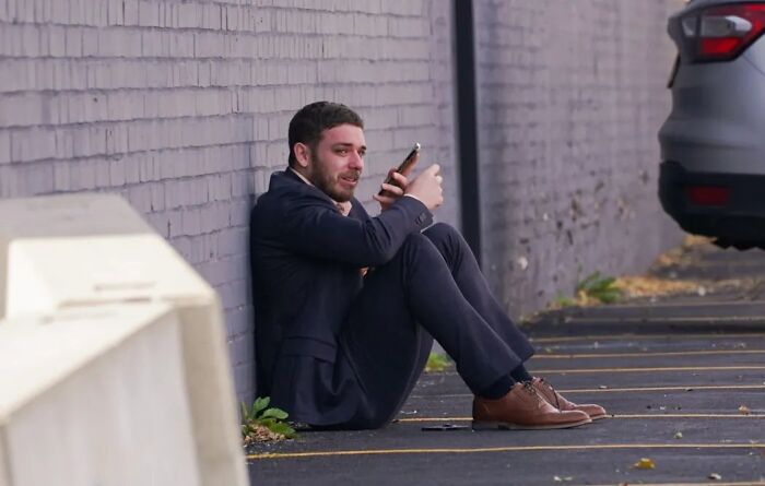 Man in tears sitting against wall outside ICE facility, holding phone after running to follow disappeared wife.