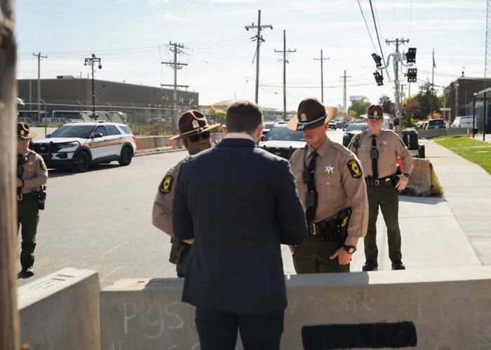 Man in tears talking to ICE officers outside detention facility on a sunny day, surrounded by law enforcement vehicles.
