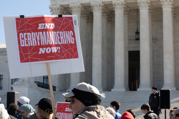 Protesters outside a government building holding signs to end gerrymandering affecting House seats ahead of 2026 midterms.