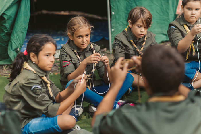 Children in Boy Scouts uniforms sitting in a circle outdoors learning knot tying during a camping activity.