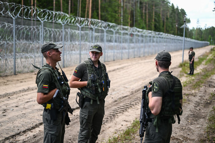 Border patrol officers in uniform standing by a barbed wire fence representing surveillance states and migration control.