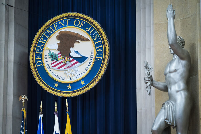 Department of Justice emblem displayed on a blue curtain backdrop beside a large statue inside a government building.