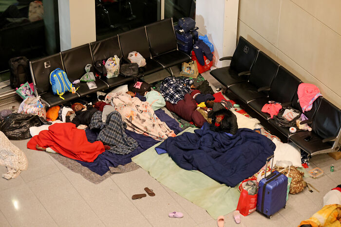 Groups of migrants sleeping on the floor and chairs inside an airport amid safety concerns over forced housing by administration.