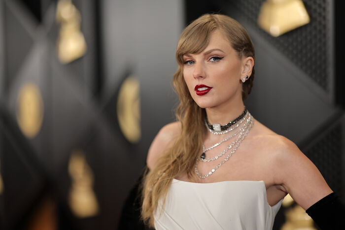 Taylor Swift at an event, wearing a white dress and layered silver necklaces, with fans outraged over music misuse.