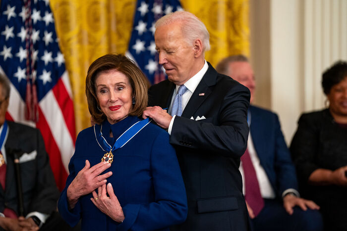 Nancy Pelosi receiving a medal from Joe Biden during a formal event with American flags in the background.