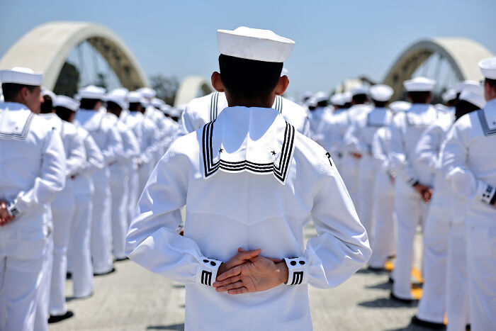 Navy sailor standing in formation wearing white uniform with sailor hat during a ceremonial event outdoors.
