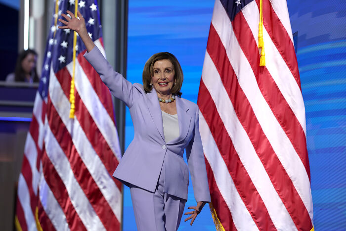 Nancy Pelosi waving on stage between American flags during announcement about not seeking re-election.
