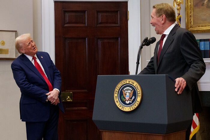 Donald Trump in a navy suit speaking with a man at a podium with the Presidential seal in a formal room