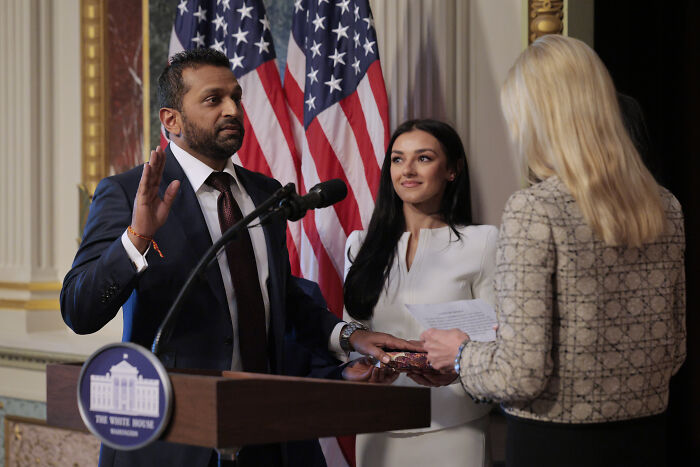 Kash Patel raising his hand at a podium with American flags in the background during a formal event.