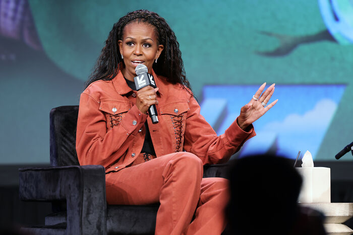 Michelle Obama speaking on stage, wearing rust-colored outfit, addressing audience about presidential run reasons.