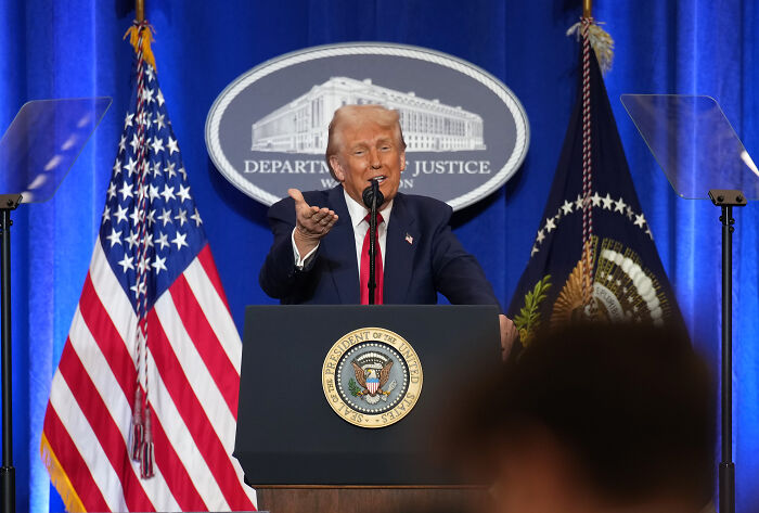 Former President Trump speaking at the Department of Justice podium with U.S. and presidential flags in background.