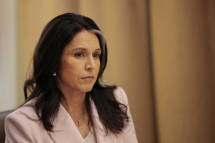 Tulsi Gabbard in a pink blazer, seated and looking thoughtful during an indoor interview or discussion.