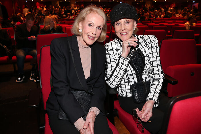 Beloved twin sisters, former entertainment icons, seated together in a theater with red seats and audience in background
