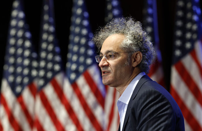 Man with curly gray hair and glasses speaking at an event with multiple American flags in the background.