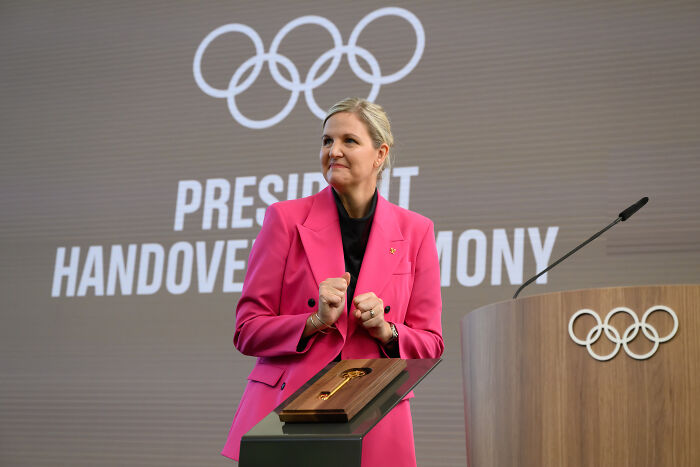 Woman in pink blazer at podium with Olympic rings during president handover ceremony for Olympics policy on transgender and DSD competitors.