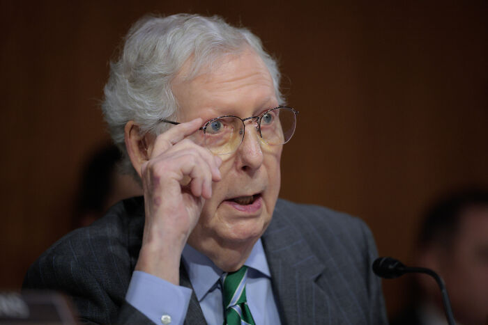 Elderly man in glasses speaking seriously at a hearing, related to government impact on family income.