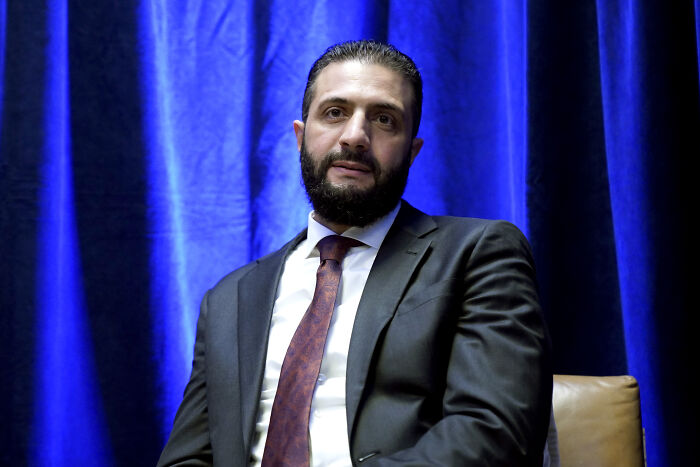 Man in suit and tie seated against blue curtain backdrop during White House meeting with Syrian president.