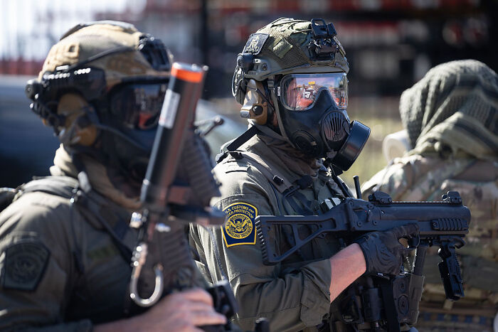 Federal agent in tactical gear holding a rifle, wearing a gas mask during a law enforcement operation.