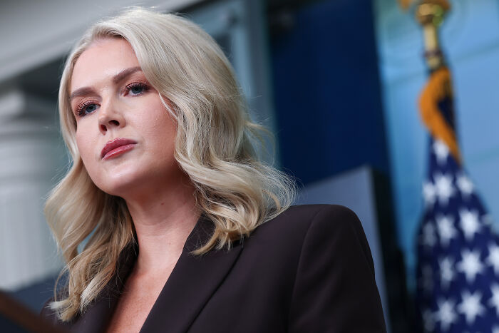 Karoline Leavitt speaking at a podium, wearing a dark blazer with an American flag in the background.