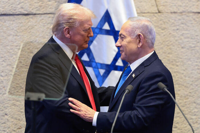 Donald Trump and Israeli leader warmly greeting each other in front of Israel flag during a formal event in 2025.