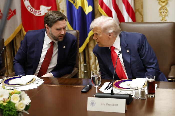 Donald Trump and JD Vance in a private conversation at a formal event with flags and a presidential nameplate visible.