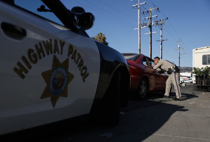 Highway patrol officer talking to driver during traffic enforcement on a sunny day near utility poles and vehicles.