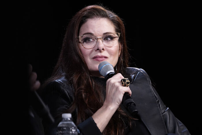 Debra Messing speaking into a microphone during an event, wearing glasses and a black leather jacket.