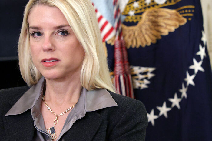Woman with blonde hair and gray blouse speaking in front of a blue and gold flag during a DOJ lawsuit discussion.