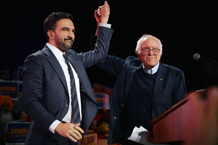 Zohran Mamdani and Bernie Sanders raising hands together at a democratic socialism event in NYC.