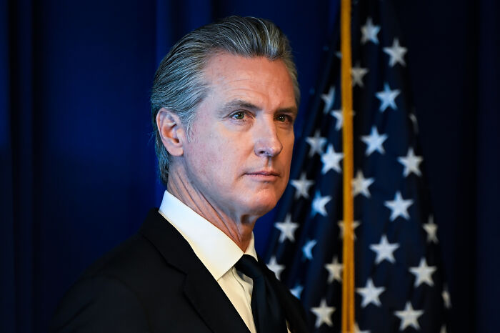 Gavin Newsom in a suit standing near an American flag, symbolizing his response after Proposition 50&rsquo;s victory.