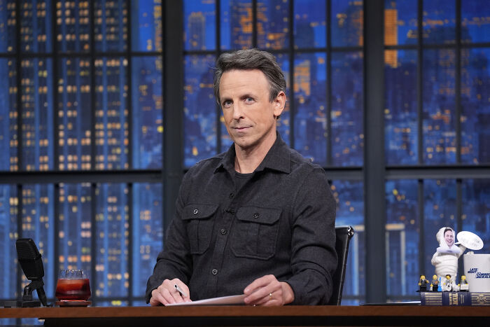 Seth Meyers seated at a desk on a late-night show set with cityscape in the background during a monologue segment.